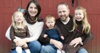 Family of five sitting together in front of a red barn, smiling and posing for a photograph.